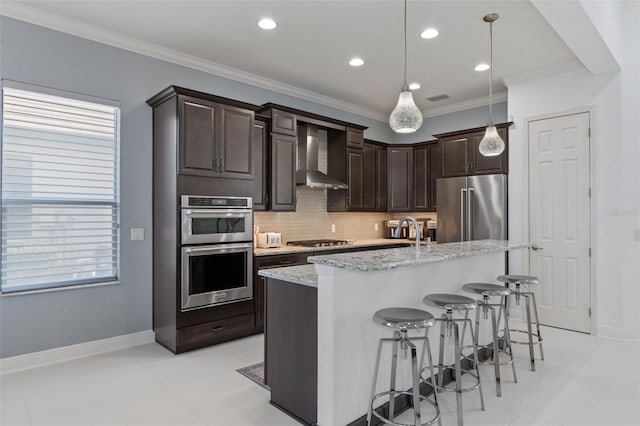 kitchen featuring an island with sink, hanging light fixtures, stainless steel appliances, light stone countertops, and wall chimney exhaust hood