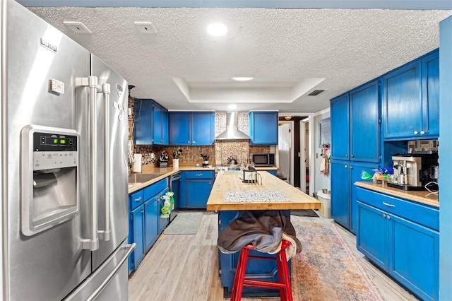 kitchen with stainless steel appliances, blue cabinetry, wall chimney exhaust hood, and light countertops