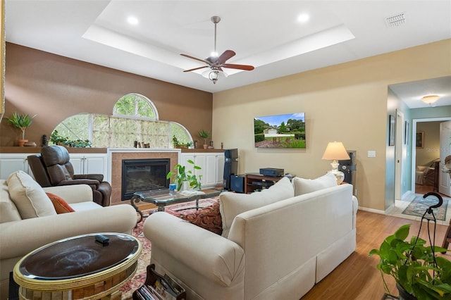 living room with ceiling fan, a fireplace, a raised ceiling, and light wood-type flooring