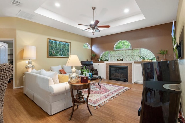 living room featuring ceiling fan, a fireplace, a raised ceiling, and light wood-type flooring