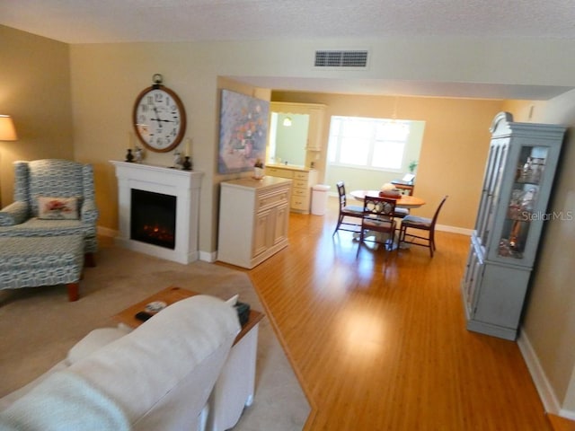 living room with visible vents, light wood-style flooring, a textured ceiling, a warm lit fireplace, and baseboards