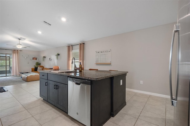 kitchen featuring sink, light tile patterned floors, appliances with stainless steel finishes, a kitchen island with sink, and dark stone countertops