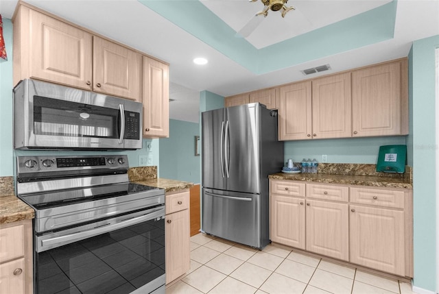 kitchen featuring light tile patterned flooring, appliances with stainless steel finishes, stone countertops, a tray ceiling, and light brown cabinets