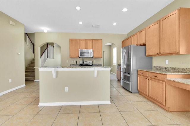 kitchen with light tile patterned flooring, a kitchen bar, light stone counters, stainless steel appliances, and a kitchen island with sink