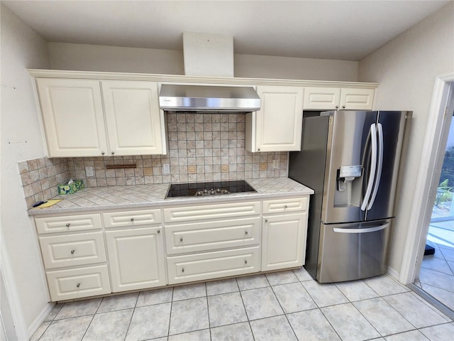 kitchen featuring tile countertops, stainless steel fridge with ice dispenser, decorative backsplash, wall chimney range hood, and black electric cooktop