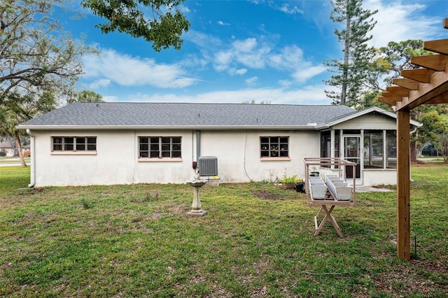 rear view of house featuring central AC, a shingled roof, a sunroom, a lawn, and stucco siding