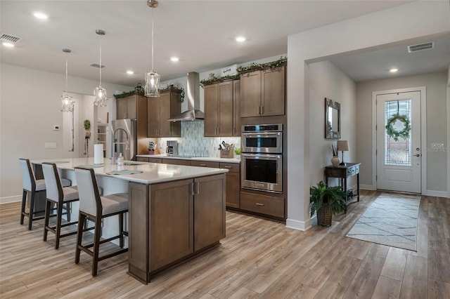 kitchen with hanging light fixtures, a kitchen island with sink, light countertops, wall chimney range hood, and a sink