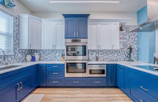 kitchen featuring stainless steel double oven, white cabinets, wall chimney range hood, light hardwood / wood-style floors, and sink