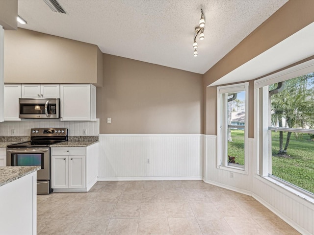 kitchen with lofted ceiling, stainless steel appliances, white cabinets, wainscoting, and light stone countertops