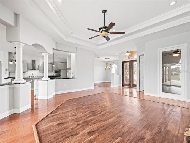 unfurnished living room with crown molding, decorative columns, a raised ceiling, wood finished floors, and ceiling fan with notable chandelier