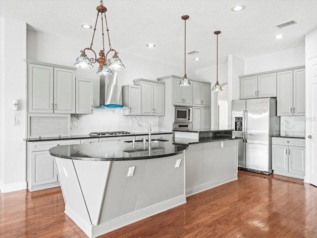 kitchen featuring decorative light fixtures, a center island with sink, appliances with stainless steel finishes, a sink, and wall chimney range hood