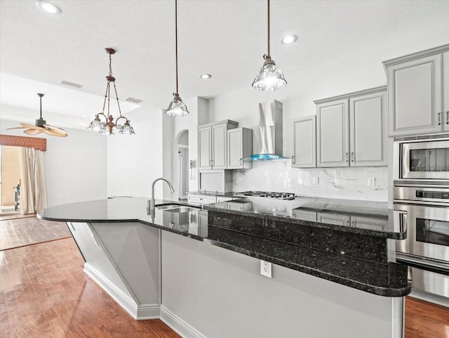 kitchen featuring hanging light fixtures, wall chimney range hood, appliances with stainless steel finishes, an island with sink, and dark stone countertops