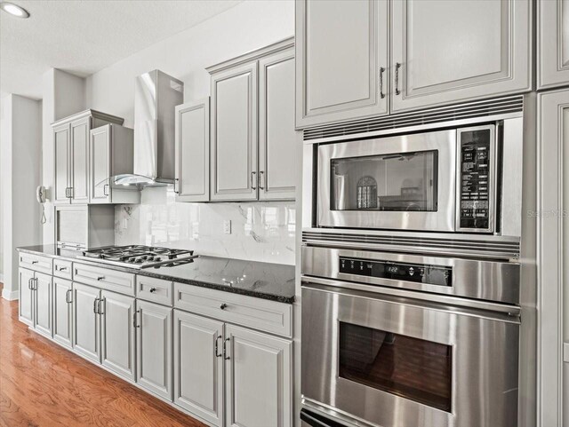 kitchen featuring appliances with stainless steel finishes, light wood-type flooring, decorative backsplash, dark stone counters, and wall chimney exhaust hood