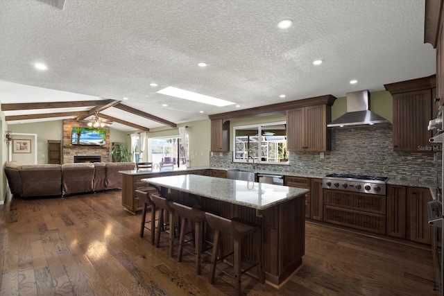 kitchen with lofted ceiling with beams, a peninsula, a warm lit fireplace, appliances with stainless steel finishes, and wall chimney range hood