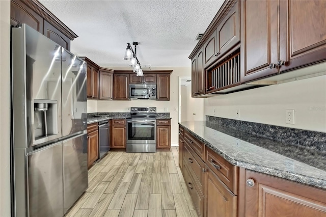 kitchen featuring a textured ceiling, stainless steel appliances, wood finish floors, dark stone countertops, and decorative light fixtures