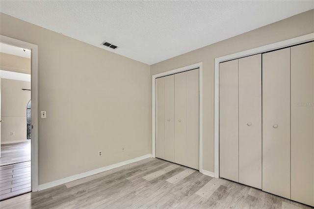 unfurnished bedroom featuring a textured ceiling, visible vents, baseboards, light wood finished floors, and two closets