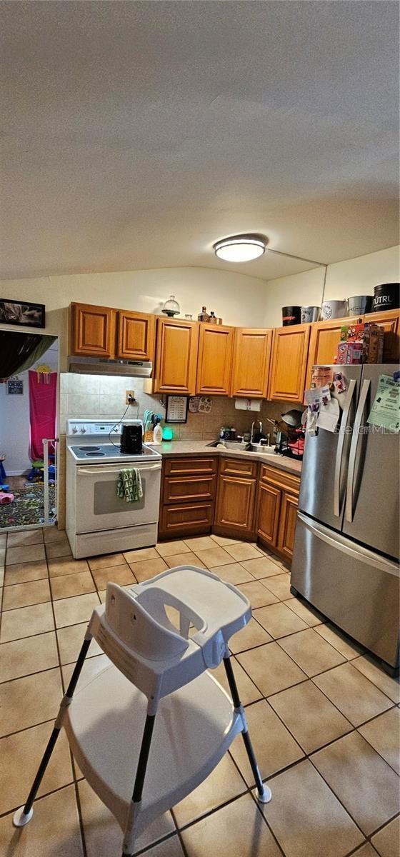 11617 Duda Rd, Hudson, FL 34667 - kitchen featuring freestanding refrigerator, electric range, under cabinet range hood, brown cabinetry, and light tile patterned floors