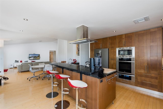 kitchen with island exhaust hood, stainless steel appliances, visible vents, light wood-style floors, and a kitchen bar