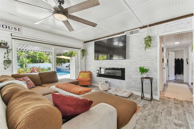 living area featuring visible vents, a glass covered fireplace, ceiling fan, brick wall, and wood finished floors