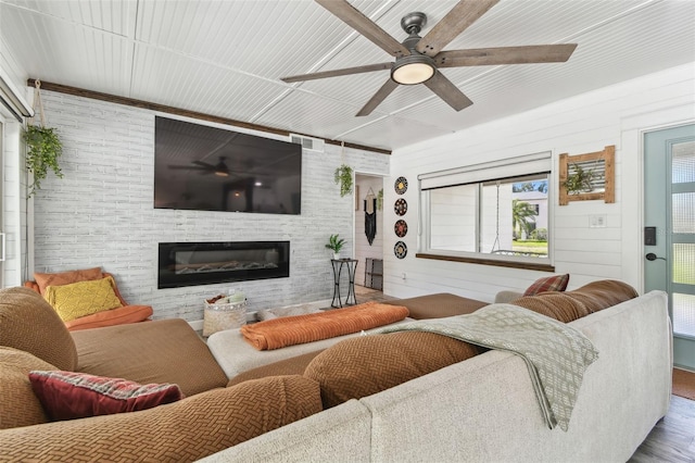 living room featuring a ceiling fan, a glass covered fireplace, visible vents, and brick wall