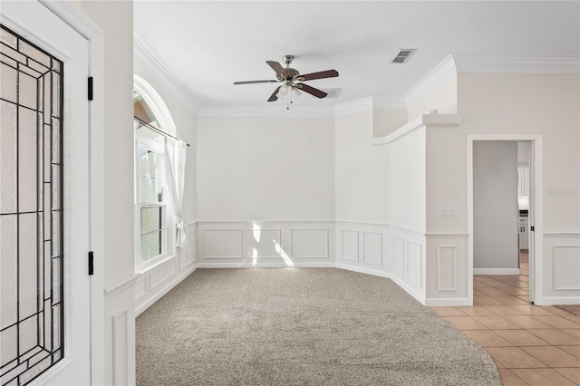 tiled spare room with ceiling fan, a decorative wall, carpet flooring, visible vents, and ornamental molding