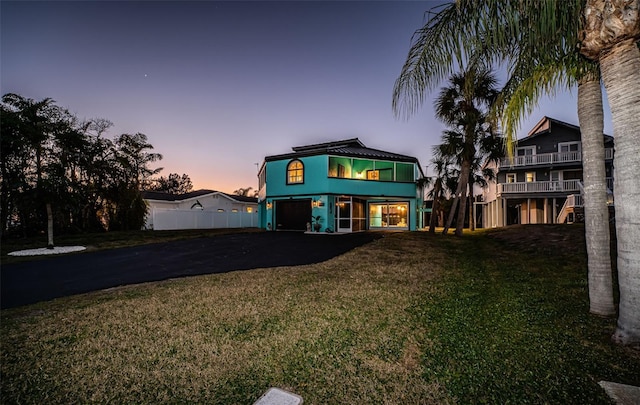 back of house at dusk featuring aphalt driveway, a balcony, a garage, fence, and a yard