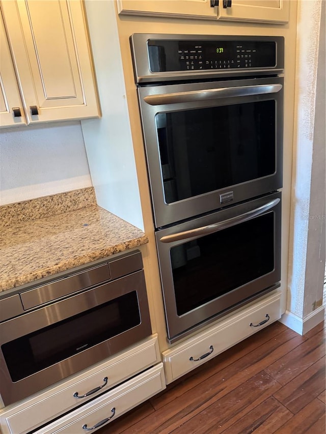 kitchen with dark wood-style flooring, stainless steel double oven, white cabinetry, light stone countertops, and baseboards