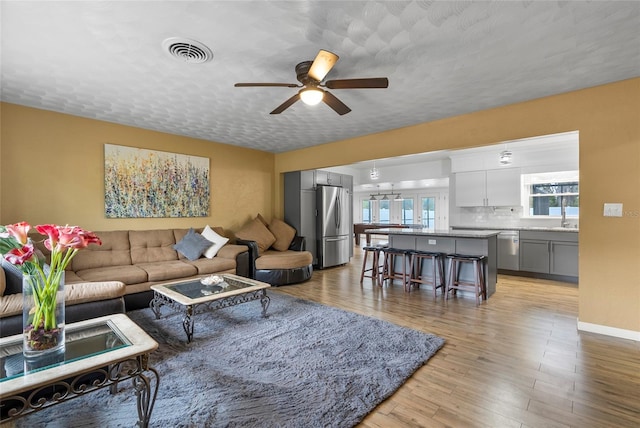 living area featuring a textured ceiling, light wood-style flooring, visible vents, baseboards, and a ceiling fan