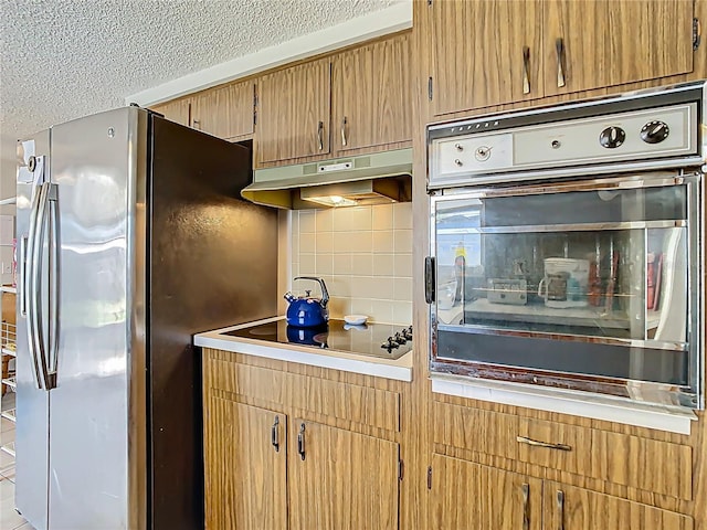 kitchen featuring black electric stovetop, oven, under cabinet range hood, backsplash, and stainless steel fridge