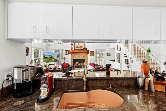 kitchen featuring a tiled fireplace, open floor plan, white cabinets, a sink, and dark stone countertops