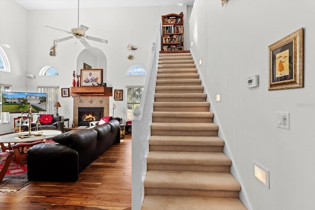 staircase featuring wood-type flooring, a high ceiling, a tiled fireplace, a ceiling fan, and baseboards