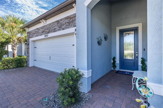 entrance to property with a garage, stone siding, decorative driveway, and stucco siding