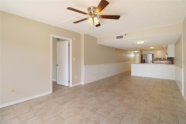 unfurnished living room featuring a wainscoted wall, a ceiling fan, visible vents, and a textured ceiling