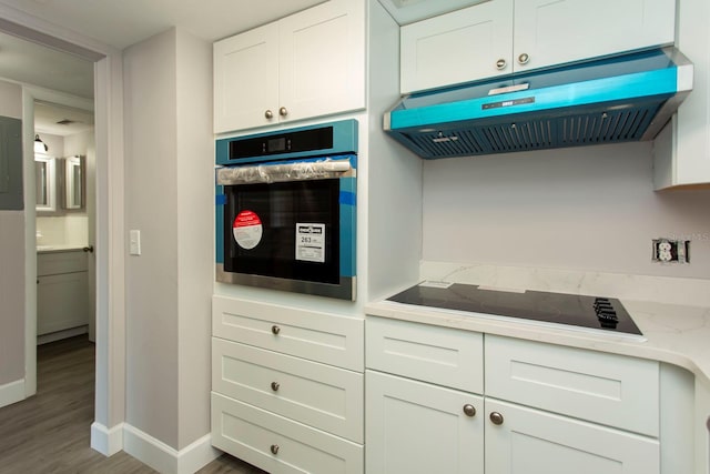 kitchen featuring oven, black electric stovetop, under cabinet range hood, white cabinetry, and baseboards