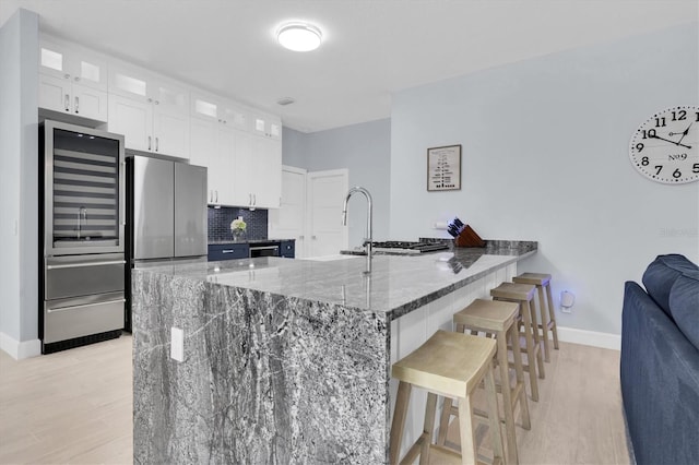 kitchen featuring glass insert cabinets, decorative backsplash, dark stone countertops, a peninsula, and freestanding refrigerator