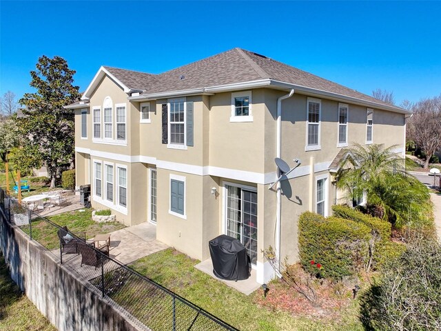 back of property with a shingled roof, stucco siding, a lawn, a fenced backyard, and a patio area