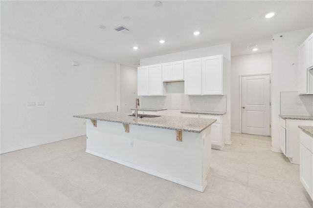 kitchen with light stone countertops, visible vents, a kitchen island with sink, a sink, and white cabinets