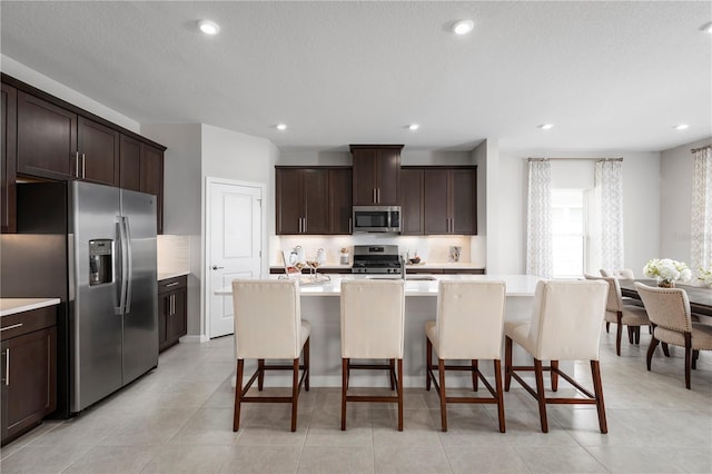 kitchen featuring an island with sink, light countertops, dark brown cabinetry, a breakfast bar area, and appliances with stainless steel finishes