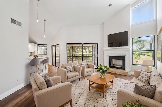 living room featuring high vaulted ceiling, wood finished floors, a fireplace with raised hearth, baseboards, and visible vents