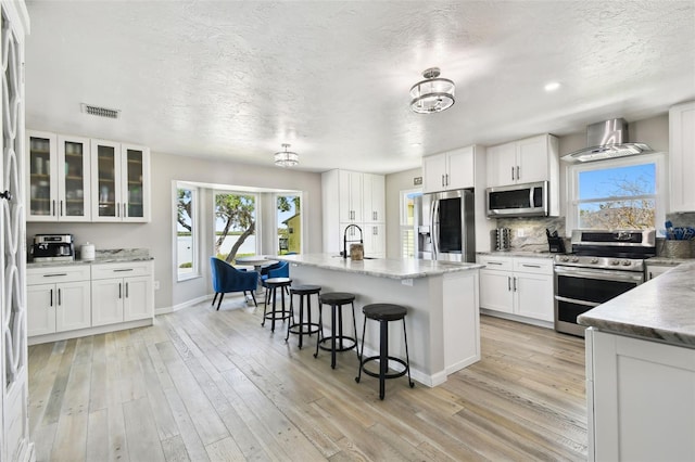 kitchen with light wood-style flooring, wall chimney exhaust hood, stainless steel appliances, white cabinets, and visible vents