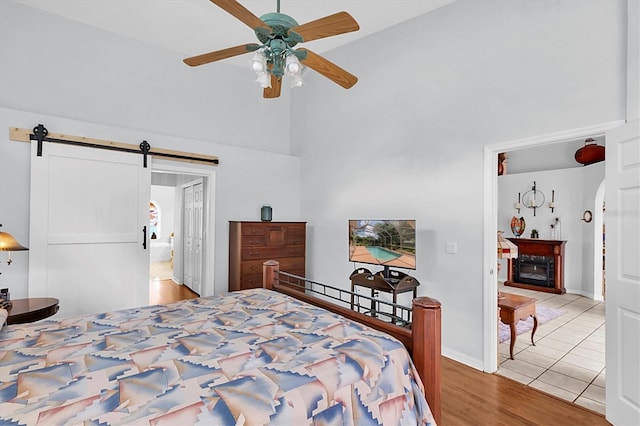 bedroom with high vaulted ceiling, a barn door, wood finished floors, a glass covered fireplace, and ceiling fan