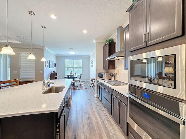 kitchen with decorative backsplash, light wood-style flooring, wall chimney exhaust hood, appliances with stainless steel finishes, and a sink