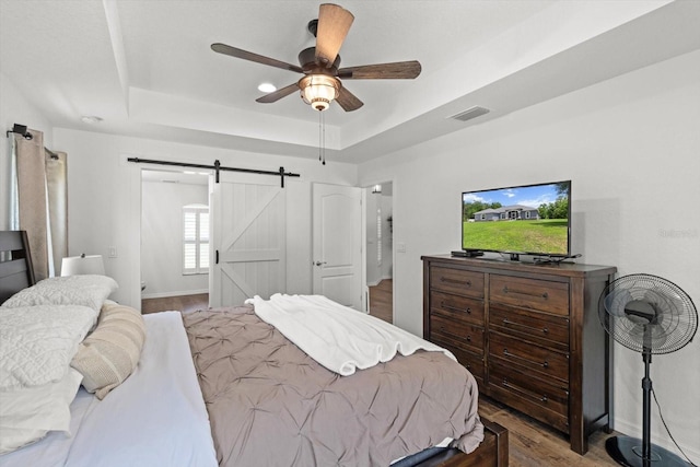 bedroom with visible vents, wood finished floors, a barn door, a tray ceiling, and a ceiling fan