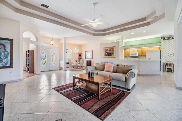 living area with a tray ceiling, visible vents, light tile patterned floors, and ceiling fan with notable chandelier