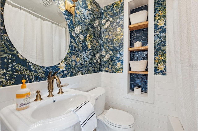 full bathroom with wainscoting, a sink, toilet, wallpapered walls, and tile walls