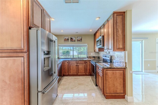 kitchen featuring brown cabinetry, backsplash, a sink, dark stone counters, and stainless steel appliances