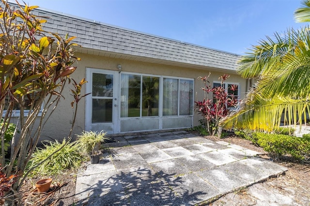 exterior space with a shingled roof, mansard roof, stucco siding, and a patio area