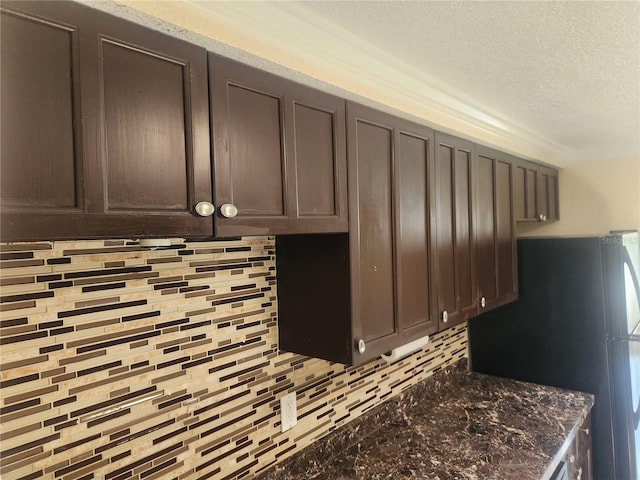 kitchen featuring freestanding refrigerator, decorative backsplash, dark brown cabinets, dark stone counters, and a textured ceiling