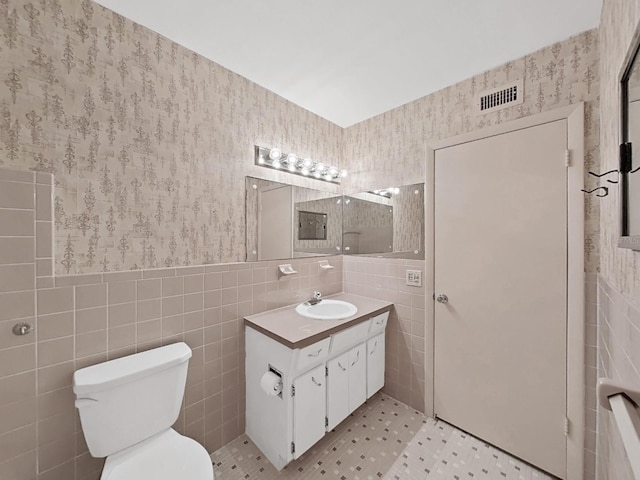 bathroom featuring tile walls, wallpapered walls, toilet, vanity, and wainscoting