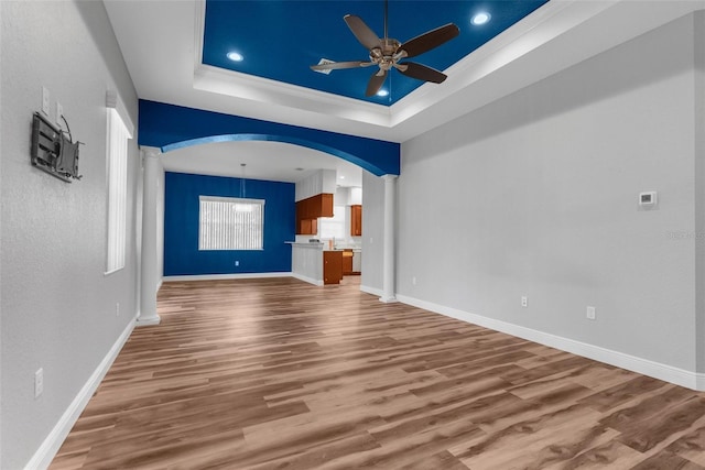 unfurnished living room with ceiling fan, a raised ceiling, wood finished floors, arched walkways, and baseboards